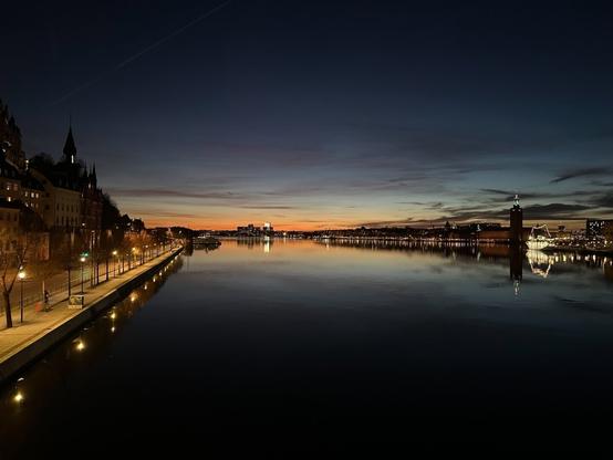 a view over water towards the last rays of the sunset. On the left, the shore stretches towards the horizon, and is lined with street lamps and trees. On the right, a brightly lit ship is moored some way in the distance.