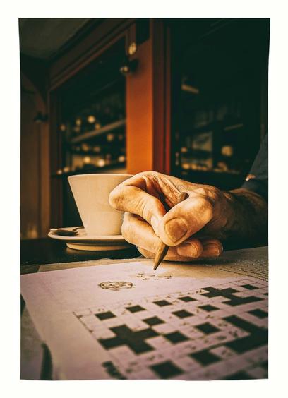 In the centre of the photo there is a man's right hand holding a pencil writing on a piece of paper. Just behind his hand is a white coffee cup. In the foreground we see the piece of paper he is writing on, it's a cryptic crossword. The background is a little dark and moody and out of focus but we can see the entrance and one of the outside windows of the café.