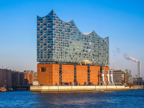 The image depicts a modern architectural building known as the Elbphilharmonie, located in Hamburg, Germany. It features a distinctive glass structure that sits atop a historical warehouse. The design includes a wavy roofline and is situated near the waterfront, showcasing both contemporary and industrial elements. The building serves as a concert hall and cultural venue, distinct for its unique design and prominence in the city's skyline.

Image Credits: Wikimedia / Hackercatxxy / CC BY-SA 4.0