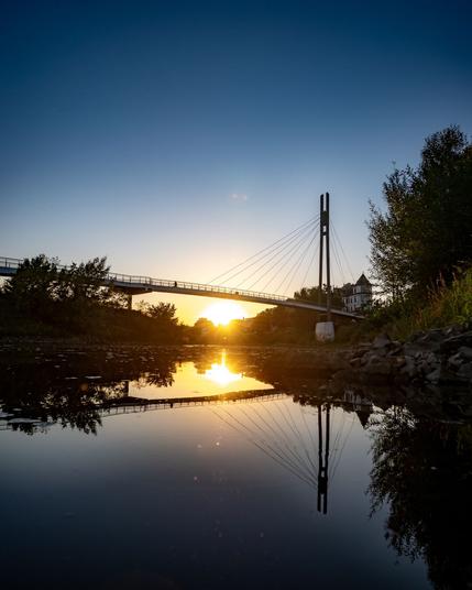 The sun is setting behind a pedestrian bridge leading over a small side arm of the main river