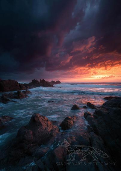A dramatic seascape at sunset, featuring jagged rocks emerging from swirling ocean waves. The sky is split between deep indigo storm clouds on the left and a fiery orange-pink sunset on the right, casting vivid reflections across the water and wet rocks in the foreground. The mood is both turbulent and serene, capturing the raw beauty of nature’s contrasts.