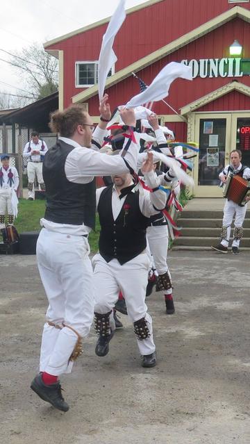 Morris dancers with white shirts and trousers and black vests, decorated with multicolor ribbons, are in front of a red building labeled "Council Rock". They are in motion, hands raised, and are in line with one another. Behind them is a musician playing an accordion and some other morris dancers.