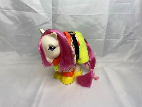 A colorful toy pony with pink hair, wearing a bright outfit featuring yellow and orange elements. The background is a plain, light-colored fabric.