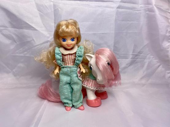 The image features a doll with blonde, curly hair wearing a turquoise and pink outfit, standing next to a toy pony with a pink mane and white body. Both are positioned against a plain white background.