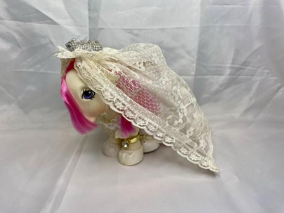 A toy pony with pink hair is dressed in a lace veil and a crown, posed on a light background.