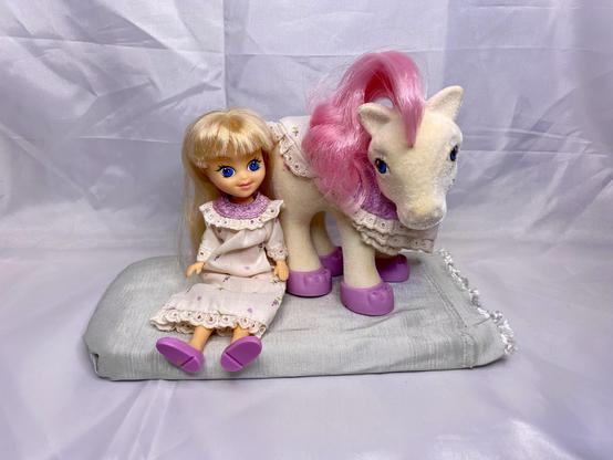 A small blonde doll wearing a white dress sits on a soft gray cushion next to a plush toy pony with pink hair and purple shoes. The background is a plain white fabric.