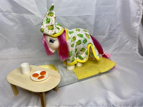 A toy pony is dressed in a patterned nightgown and hat. It is positioned on a soft surface near a small wooden table that holds a white cup and a plate with three orange cookies. The background is a simple, light fabric.
