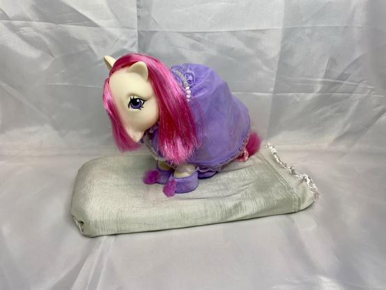 A toy pony with pink hair and a purple outfit is positioned on a light gray fabric. The pony is looking downward and sits on a bed. The background is a smooth, light-colored surface.