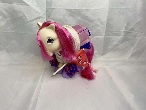 A colorful pony toy with long pink hair, dressed in a white shirt and purple skirt, holding a small flag labeled "MLP." The background is plain and light-colored.