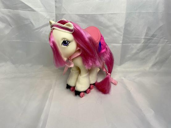 A toy horse with pink hair is wearing roller skates and a saddle. The horse has large eyes and is posed on a white background.