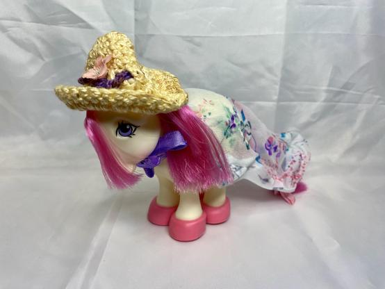 A toy horse with pink hair, wearing a straw hat and a floral dress, stands against a plain white background.