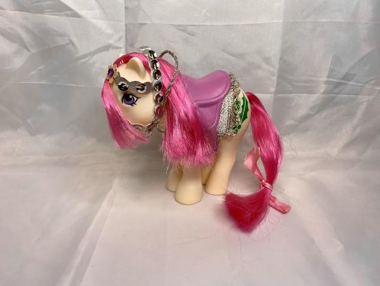 A toy pony with long pink hair, adorned with a silver chain and headpiece, wearing a purple saddle and a decorative outfit. The background is plain and white.