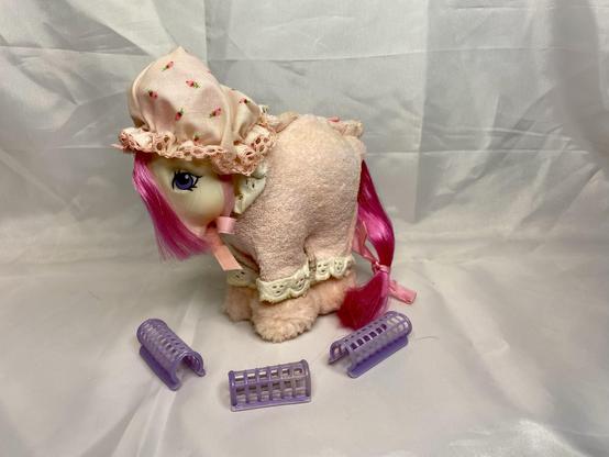 A plush toy pony with pink hair wearing a bonnet and a dress, accompanied by three small purple hair curlers on a light background.