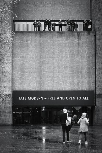 Black and white. A man and a woman are walking towards the Tate Modern art gallery. It's a very tall brick-built former power station. There is a wide entrance with the words, "Tate Modern - Free and open to all" above it. High up, near the top of the picture is a balcony with a chest-height metal railing where eleven people are looking out at the view. One is currently looking at his phone, possibly researching St Paul's Cathedral which is almost opposite, across the River Thames.