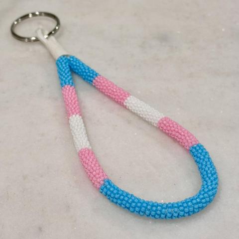 A bead wrapped keychain in the trans pride colors on a white marble background