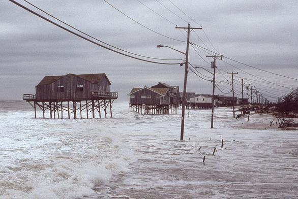 The view is west looking into the distance down a road that is hidden by crashing waves from the receding "no-name" storm of October 1991 that later became known as the "perfect storm" for the confluence of meteorlogical factors that caused it. Houses on stilts, on after another, are seen undermined by frothing waves. The sky is a heavy, leaden gray. Utility poles and their wires are still standing.