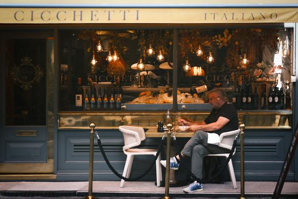 A man is sitting at an al fresco dining table outside a San Carlo Italian cicchetti restaurant in central London. The window behind him is full of seafood and wine with dried leaves, hanging fruit, and eleven lit lightbulbs above. Cicchetti is more or less the Italian version of tapas, and is usually served on small plates. Perhaps he's having a dolce, or dessert.