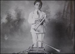Antique vaudeville promo photo of Galla-Rini at age 9 or so in a neat little pale suit, holding a cornet. At his feet are other horns and a button accordion.
