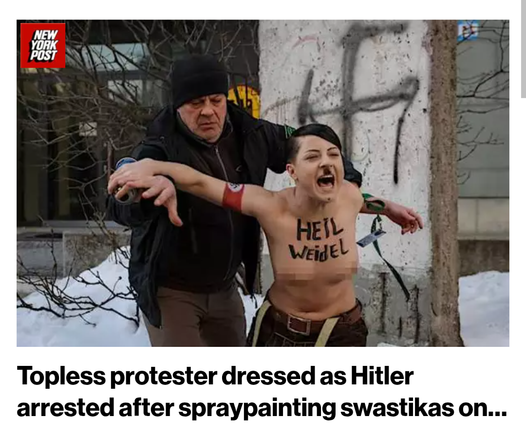 Topless woman with Heil Weidel pained on her chest, a Hitler mustache painted on her upper lip, and an Nazi armband pained on her upper arm screams as she is arrested by an older man in a jacket and beanie cap. There is a swastika on the ruins of the Berlin wall behind them. There is snow on the ground.