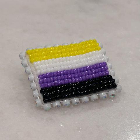 A beaded pride flag pin in the nonbinary pride colors -- yellow, white, purple, and black -- on a white marble background. The pin is edged with glass rhinestone banding.