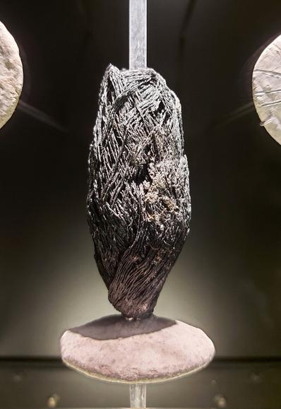 A charred skein of linen thread displayed upright in a museum case. The blackened, twisted fibers are tightly coiled and partially fused from burning, mounted between two smooth, pale stone supports. The exhibit is lit to highlight the texture and preservation of the  textile remains.