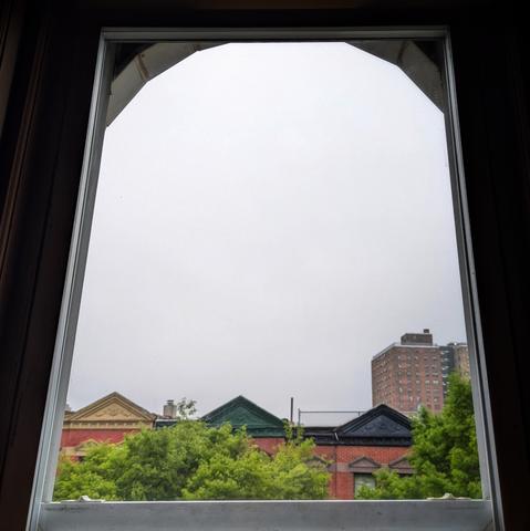 Looking through an arched window an hour after sunrise the sky is full of grey fog. Pointed roofs of Harlem brownstones with red brickwork are across the street, and a taller apartment building can be seen in the distance. The green tops of two trees are on the bottom and right.