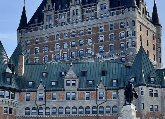 A large multi-tiered chateau with rows and rows of windows. The foreground has a human statue (black on a beige stone pedestal) in front of a building with long rows of rectangular and rounded windows, including some set into a sloped green copper roof. In the background above is an older building with rows of rectangular windows inset in beige and bien brick. A light blue sky with wispy white clouds fills in behind. [Le Château Frontenac (PQ, Canada), May 2025]