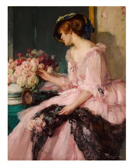 A red haired woman wearing a hat and a pink dress sits and arranges flowers in a vase.