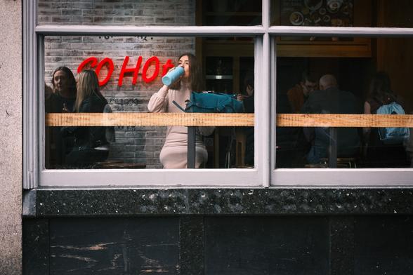 A young woman with brown hair, dressed in a tight, high-necked woollen dress is sitting on a stool in the window of a Soho Coffee bar, drinking out of a long pale blue flask. Her flask, her blue bag on the breakfast counter, and the blue backpack of another customer, suggest the idea of coolness. In contrast are the words, "So Hot" on the wall behind the woman, in an almost scarlet red, with glowing pink light emanating from behind them.