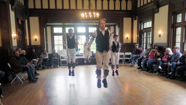 Three morris dancers with white shirts and trousers and black vests, decorated with multicolor ribbons, are in a large Tudor revival room. All are off the ground with hands down and behind them. Behind them is a morris dancer in white. To the sides are audience members in chairs.