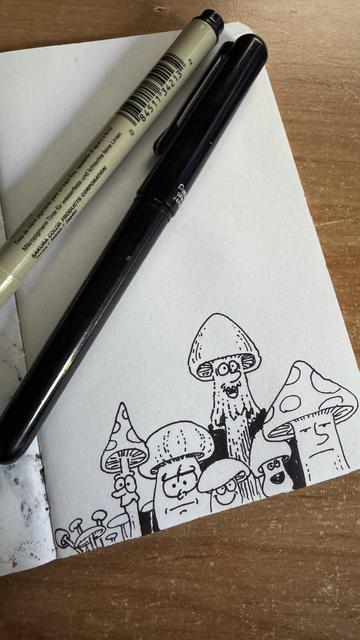Photo of a sketchbook page with some doodles of mushrooms with faces in different shapes. On top of the page there are a fineliner marker and a pen brush.