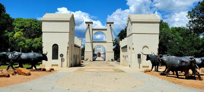 Wide view of the beige towers at the entrance to a suspension bridge with green trees behind the towers and sculptures of cattle rushing to enter the bridge. Blue skies with some white clouds above the bridge.