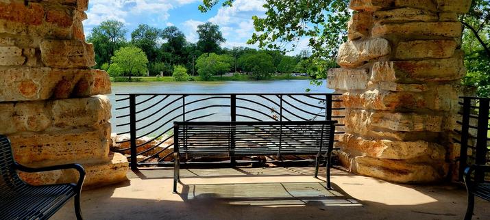 View from behind of the inside of a pavilion by a river. Black metal park bench with backrest facing a matching railing, both between yellow-orange stone pillars. The bench and the railing are sunlit. River on the other side of the pavilion, green tree line and banks on the far side.