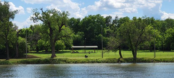 Green tree line and riverbank on the far side of a river. A pavilion with picnic benches on the far side.
