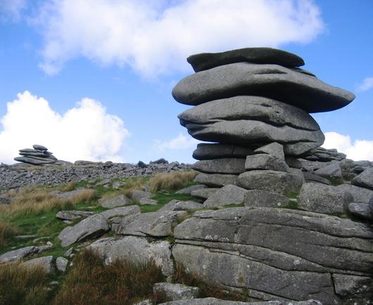 A pile of rock slabs that appear to be placed one on top of the other but are actually naturally formed. In the background is a similar structure. The sky is blue with fluffy clouds.