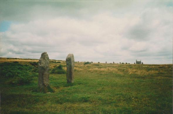 Colour photograph showing a close up view of, on the left, a pair of prehistoric standing stones or menhirs in Cornwall called The Pipers, surrounded ny the moors. Part of the three stone circles of The Hurlers can be seen on the horizon to the right-centre, with an old engine house or similar in the far distance to the right. The sky is blue and full of clouds.