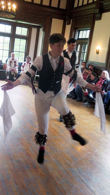 Two morris dancers with white shirts and trousers and black vests, decorated with multicolor ribbons, are in a large Tudor revival room. They are in motion, left legs raised and arms out at sides. Behind them are two morris dancers in white. To the right side are audience members in chairs.