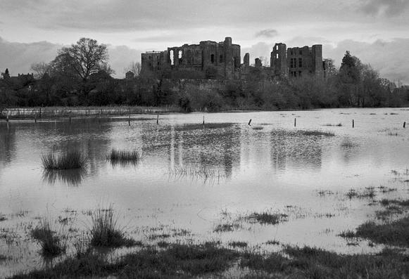 Black and white photo showing a ruined castle on the skyline, partly reflected in a flooded field. The poles of a fence can be seen with just the tops emerging from the water, which is rippled by a  slight breeze. Several clumps of reeds on the near left side.