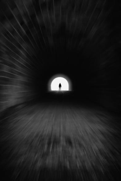 Black and white. portrait format. A long pedestrian tunnel. gloomy, cramped atmosphere. at the end of the tunnel, glistening daylight. in the light, the black silhouette of a woman standing firm and strong at the end of the tunnel. the central perspective and an added zoom effect give the picture an incredibly strong dynamic despite the gloom.