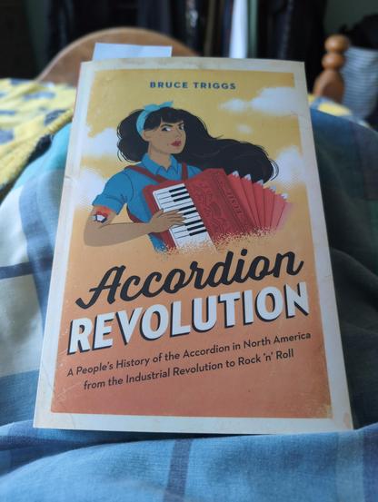 A colourful paperback book by Bruce Triggs with a cartoon of a determined looking woman on the cover playing a red piano accordion with red straps. The sleeves of her blue shirt are rolled up revealing a flaming accordion tattoo. She has long flowing black hair tied with a blue ribbon. The title of the book is Accordion Revolution - A people's history of the accordion in North America from the Industrial Revolution to Rock 'n' Roll.