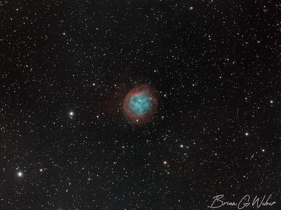 A planetary nebula in the middle of the frame with a red ring around the outside, a teal center, and faint dust lines throughout.