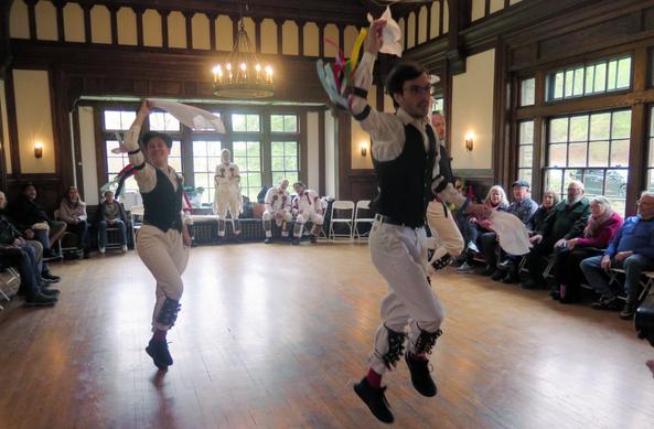 Three morris dancers with white shirts and trousers and black vests, decorated with multicolor ribbons, are in a large Tudor revival room. All are in motion with right legs and arms raised. Behind them is are three morris dancers in white. To the sides are audience members in chairs.