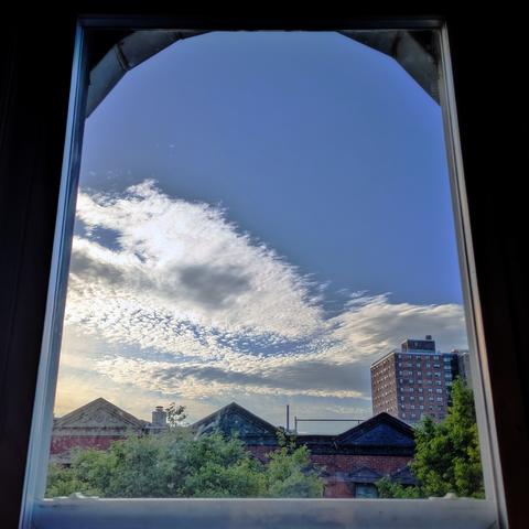 Looking through an arched window ninety minutes after sunrise the bright blue sky is half-full of spiky fluffy white clouds. Pointed roofs of Harlem brownstones with red brickwork are across the street, and a taller apartment building can be seen in the distance. The green tops of two trees are on the bottom and right.