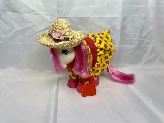 A toy horse with long pink hair, wearing a straw hat adorned with a flower and a yellow dress featuring pink flowers. It is holding a small red handbag. The background is a light gray fabric.