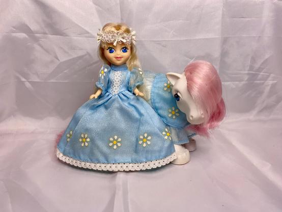 A doll in a blue dress with floral designs sits beside a white pony with a pink mane. The background is a light, neutral color.