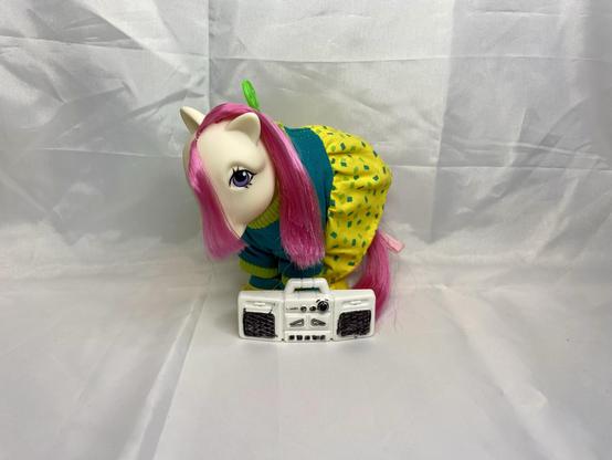 A colorful toy pony with pink hair, wearing a blue and yellow outfit, stands beside a small white boombox on a plain background.