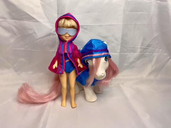 A fashion doll wearing a pink and blue hooded outfit and sunglasses stands next to a white pony adorned with a matching blue hood. Both have pink hair accents, set against a light background.