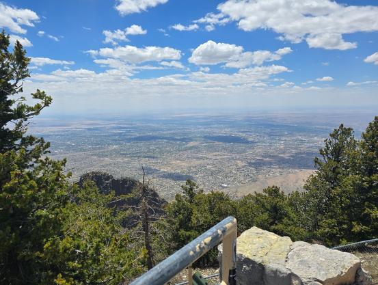 Panoramic view of Albuquerque NM from the Sandia Crest at an elevation of 10,679 feet (3,255 m). City elevation is 5312 feet (1619 m), 7424 ft (2'263 m) below. Albuquerque is relatively flat with few tall buildings or large lakes. In the foreground are pine trees that grow in the Sandia mountains. Typical rainfall is 9 inches  (24 cm) per year.