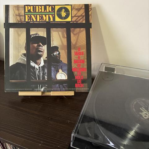 The image features a vinyl record of Public Enemy's album "It Takes a Nation of Millions to Hold Us Back," displayed me t to a turntable playing the record. The album cover shows the group’s two rappers behind bars. Flavor Flav is wearing a huge clock.