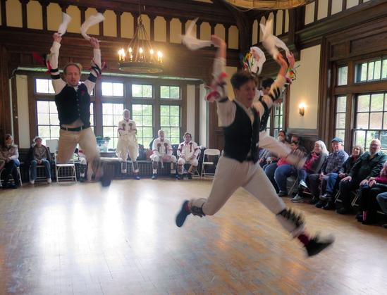 Three morris dancers with white shirts and trousers and black vests, decorated with multicolor ribbons, are in a large Tudor revival room. All are in blurred motion, leaping off the ground with both hands high. Behind them is are four morris dancers in white. To the sides are audience members in chairs.